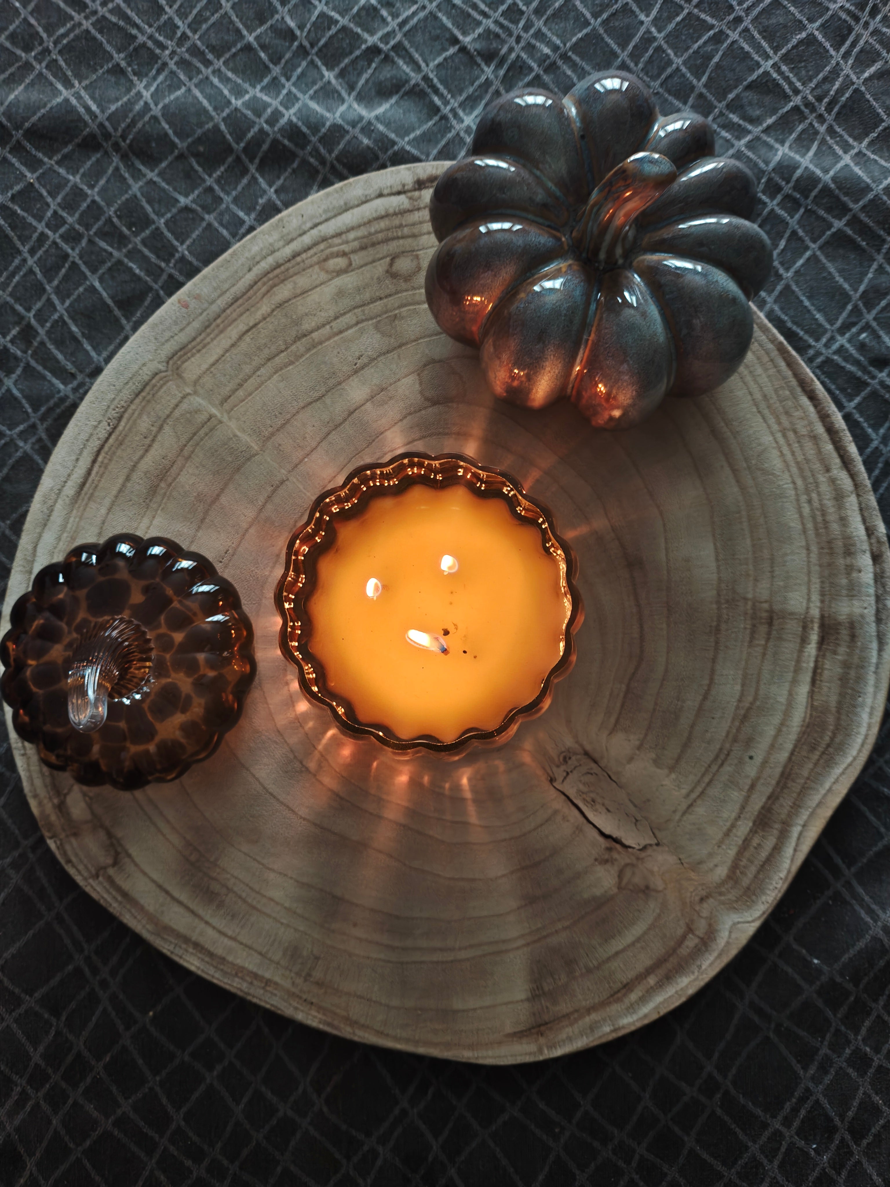 A lit candle in an amber leopard print pumpkin glass jar with matching lid on the side, styled on a round wood slice with a dark metallic pumpkin-shaped ornament on a dark textured fabric surface.