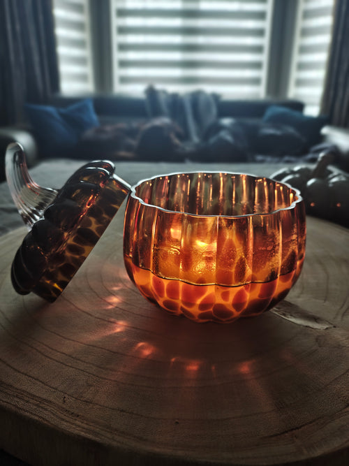 An amber leopard print pumpkin shaped glass candle jar with a matching lid, placed on a wooden surface in a dimly lit room. The bowl has a ribbed, textured exterior and is illuminated from within, casting a warm orange glow onto the wood and floor.