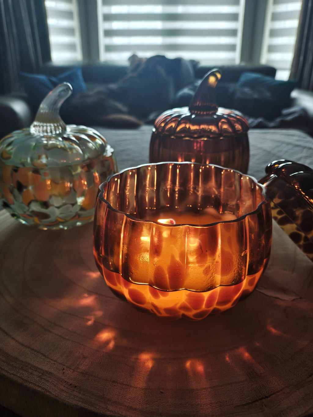 A lit amber leopard print pumpkin shaped glass candle jar sits on a wooden table in a home setting, casting a warm glow and shadow. Other decorative glass pumpkins candle jars are visible in the background. 