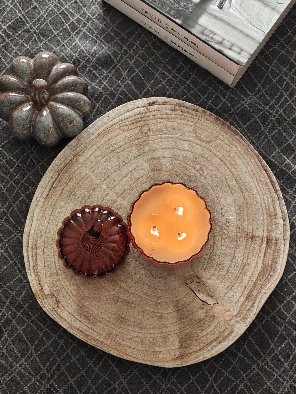 A styled flat-lay featuring a large natural wood slice tray holding a lit amber-colored pumpkin shaped candle and a matching lid on the side, set on a dark gray patterned surface with a small decorative pumpkin and a book edge visible. 