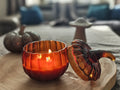 An amber glass pumpkin-shaped candle holder with a lid, shown lit on a wooden tray in a cozy indoor setting.