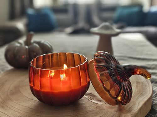 An amber glass pumpkin-shaped candle holder with a lid, shown lit on a wooden tray in a cozy indoor setting.