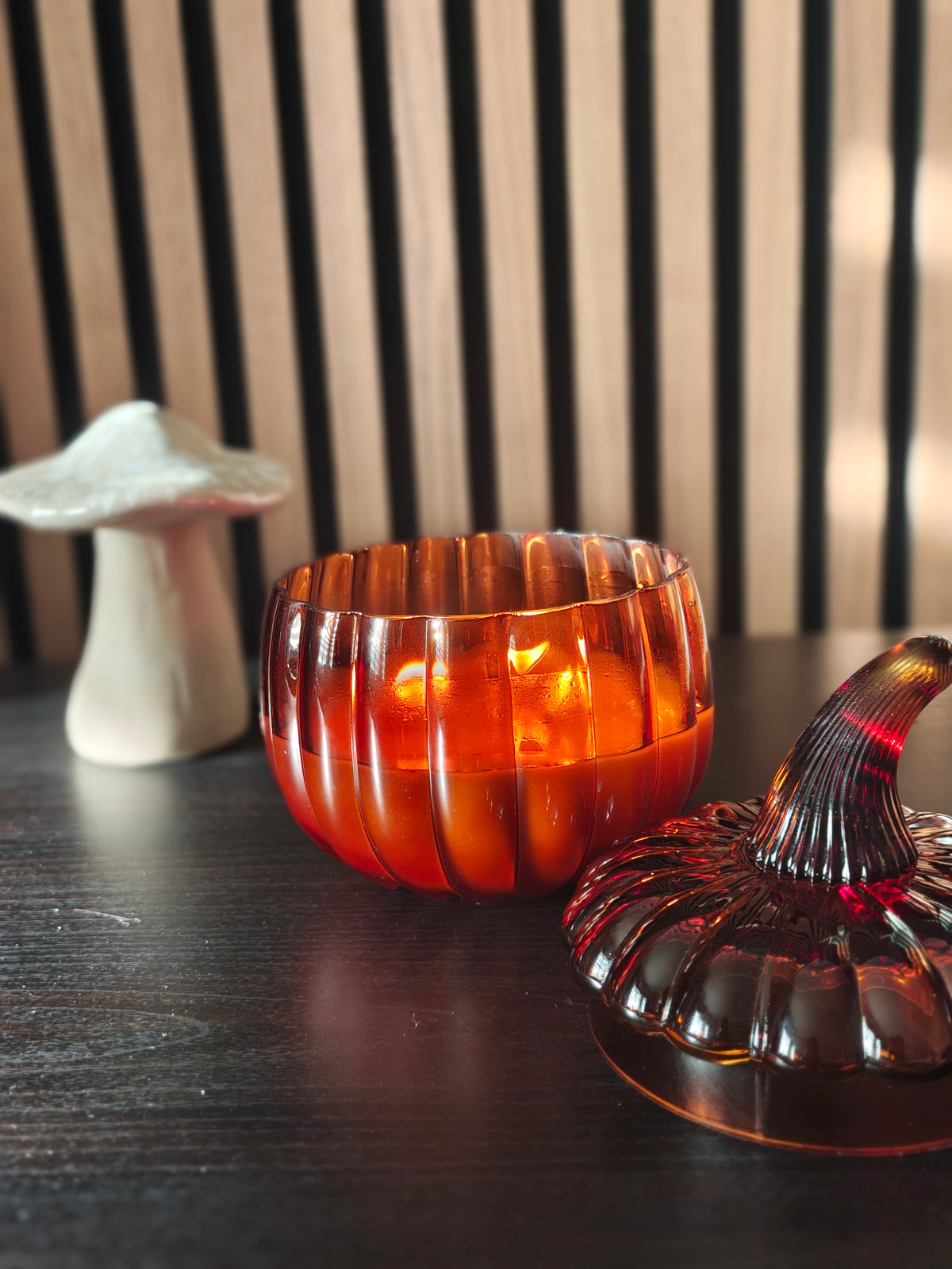 An amber-orange ribbed glass pumpkin-shaped candle holder with a matching dark glass pumpkin topper, shown on a dark wooden surface next to a white mushroom-shaped ornament.