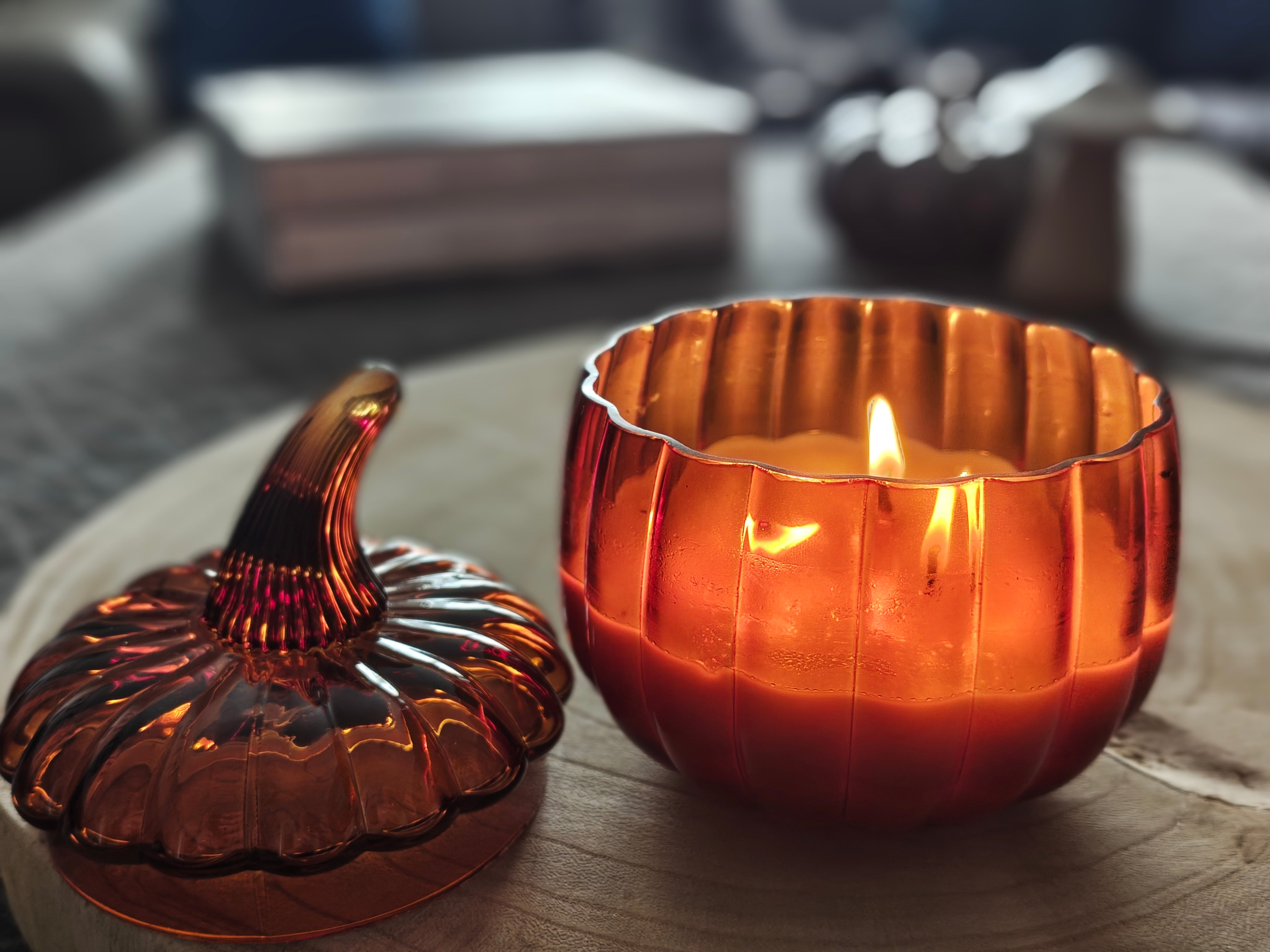An amber glass pumpkin-shaped candle holder with a lit candle inside, shown next to its matching lid on a wooden surface in a softly blurred indoor setting.