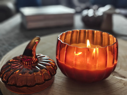 An amber glass pumpkin-shaped candle holder with a lit candle inside, shown next to its matching lid on a wooden surface in a softly blurred indoor setting.