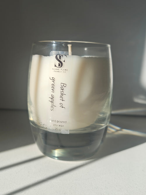 Candle in a glass holder labeled 'Basket of green apples' on a white sunlit surface.