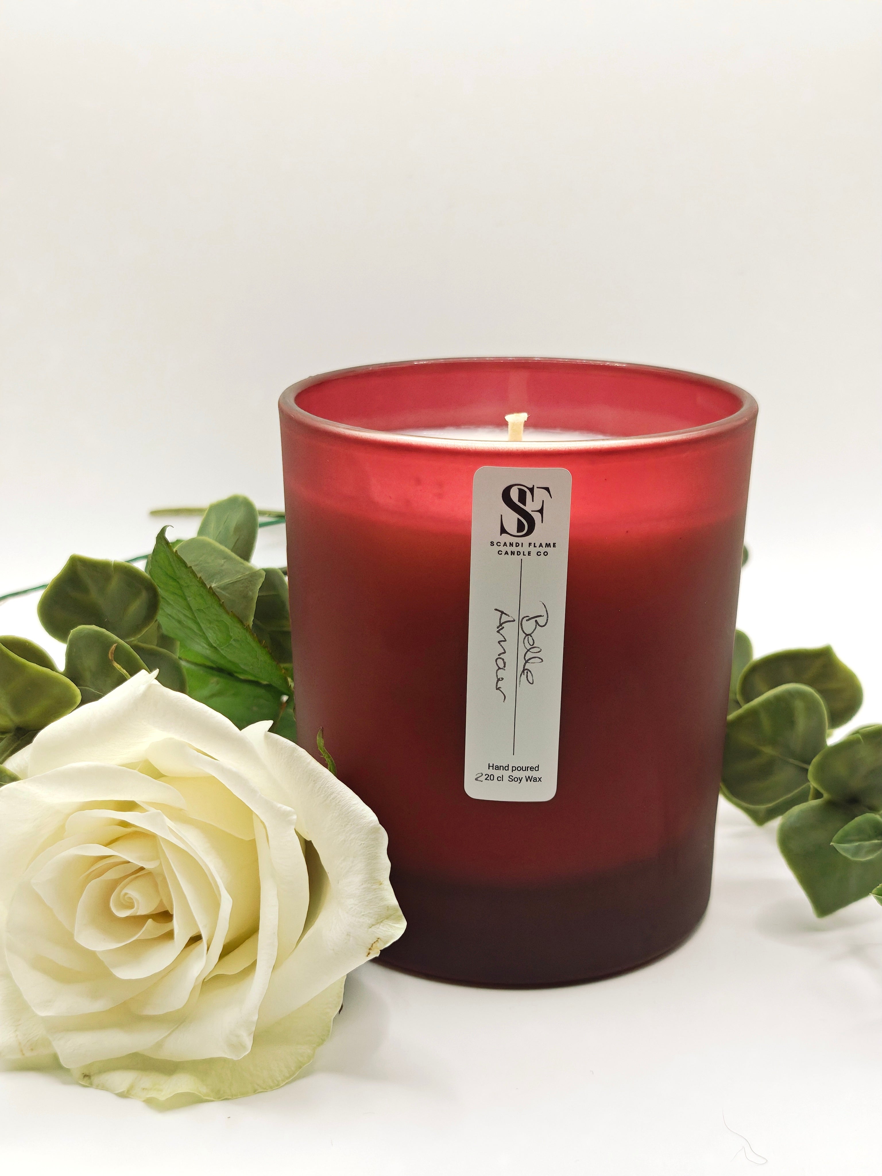 Red candle with a label on a white surface with a white rose and green leaves.