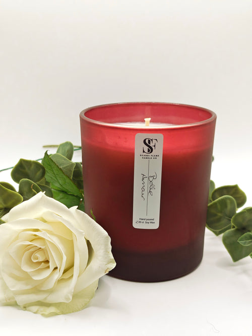 Red candle with a label on a white surface with a white rose and green leaves.