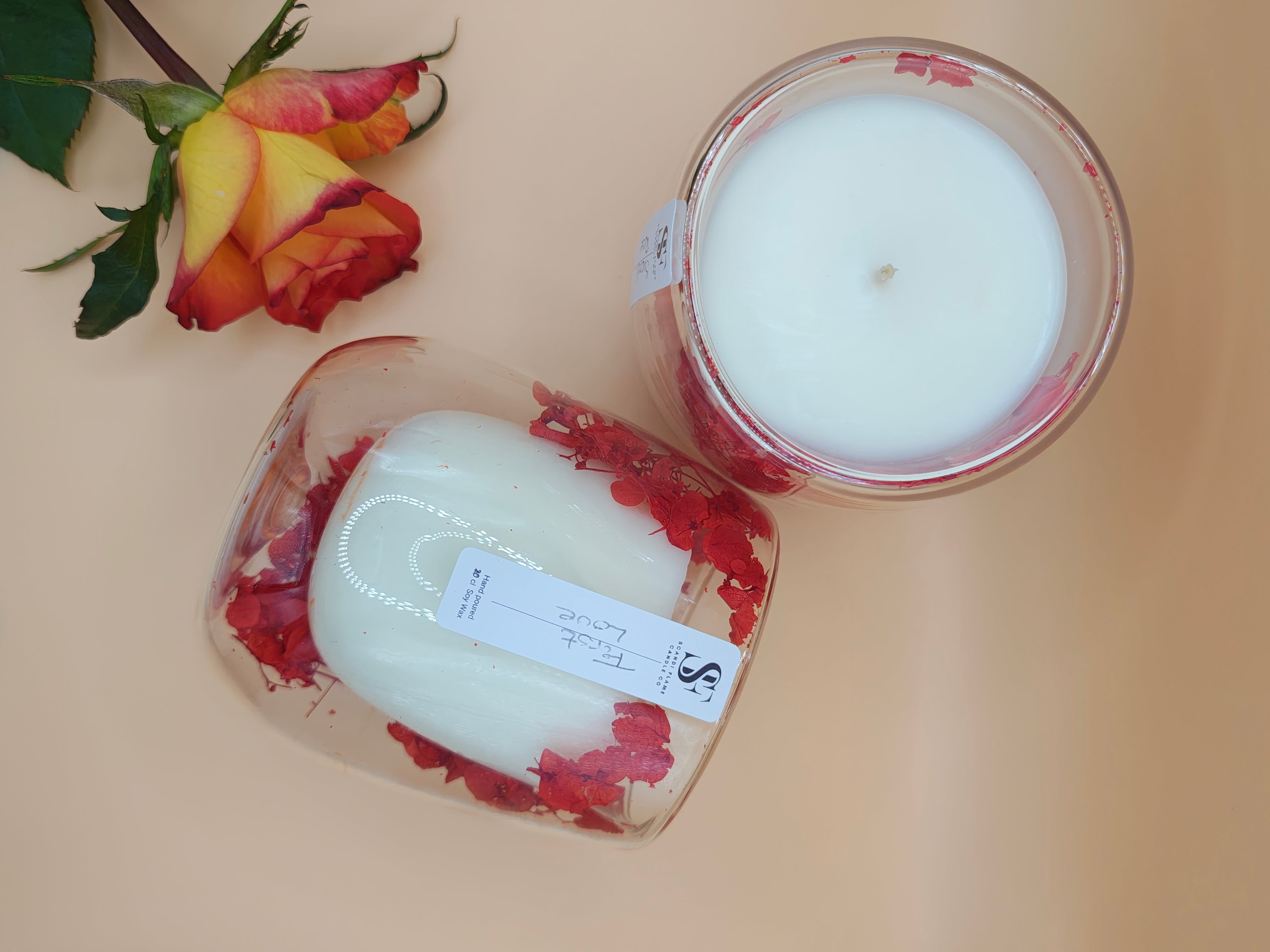 Clear glass jar with red dry flowers in the walls of the jar with 'First Love' label from Scandi Flame with a rose in the background and one jar laying down