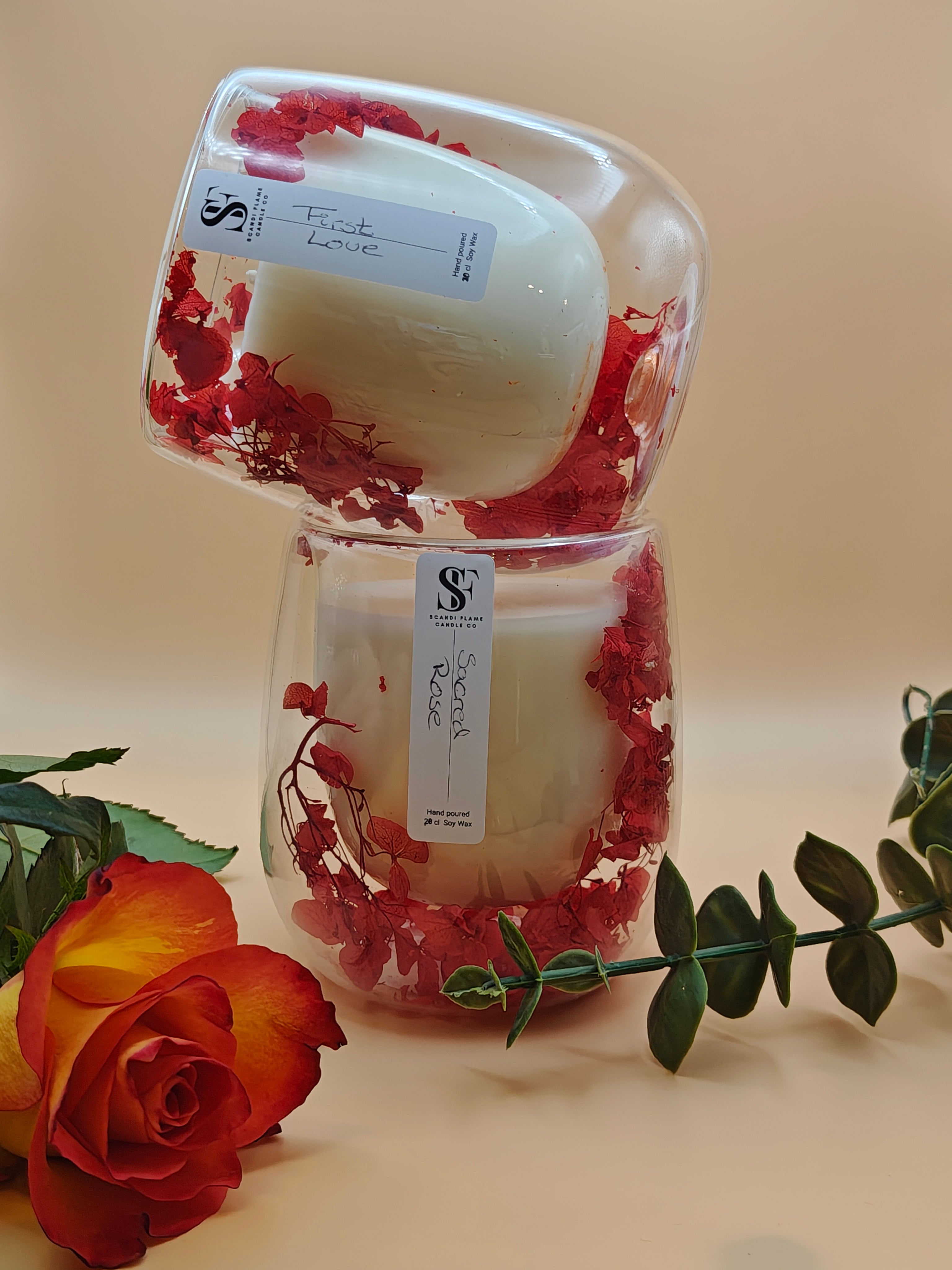 2 clear glass jar with red dry flowers in the walls of the jar. One on top of the other with 'First Love' & 'Sacred Rose' label from Scandi Flame with a rose in the foreground