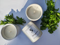 Two candles and a container labelled 'Herb Garden' on a white surface surrounded by fresh herbs.