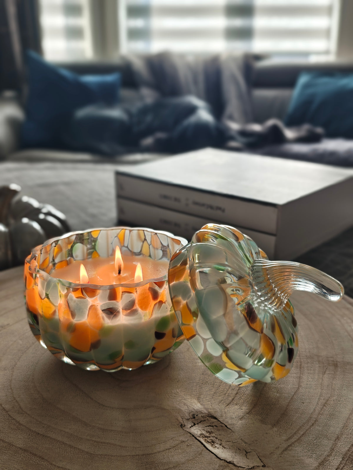 A decorative glass pumpkin-shaped candle holder with a lid, featuring a warm orange glow from internal candles, set against a rustic wood background.