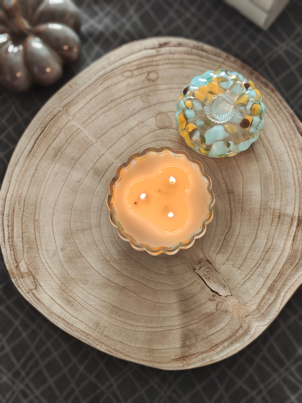 A scented candle in a warm leopard print pumpkin shaped glass jar pictured from above, sitting on a round natural wood slice, with a matching decorative lid on the side. A pumpkin ornament placed beside it on a dark patterned surface.