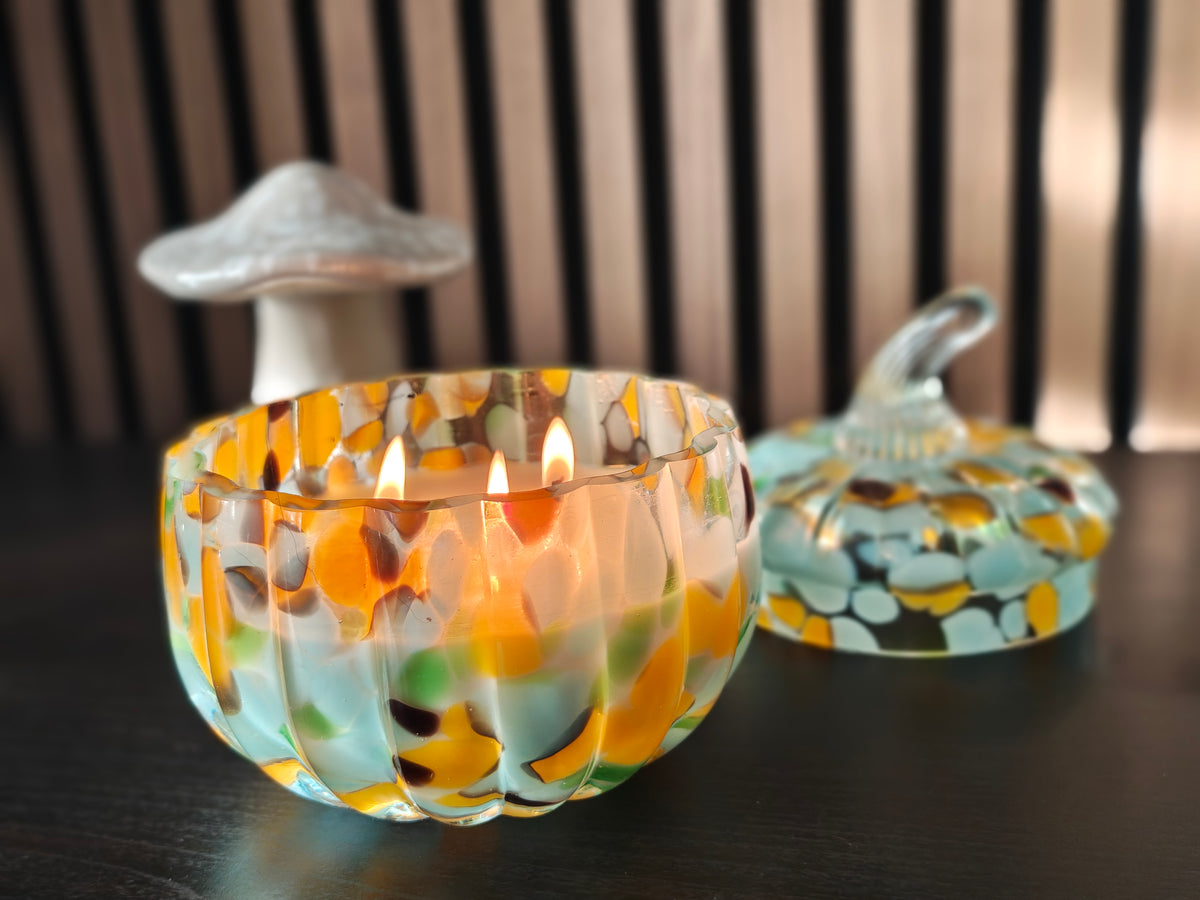 A glass candle holder with a multicolor spotted pattern in orange, yellow, green, and blue, shown lit on a dark table with a matching lid in the background.
